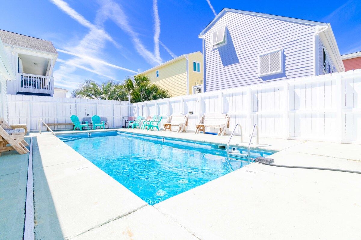 Une piscine entourée d’une clôture blanche, une terrasse en bois et une petite maison dotée d’un balcon.