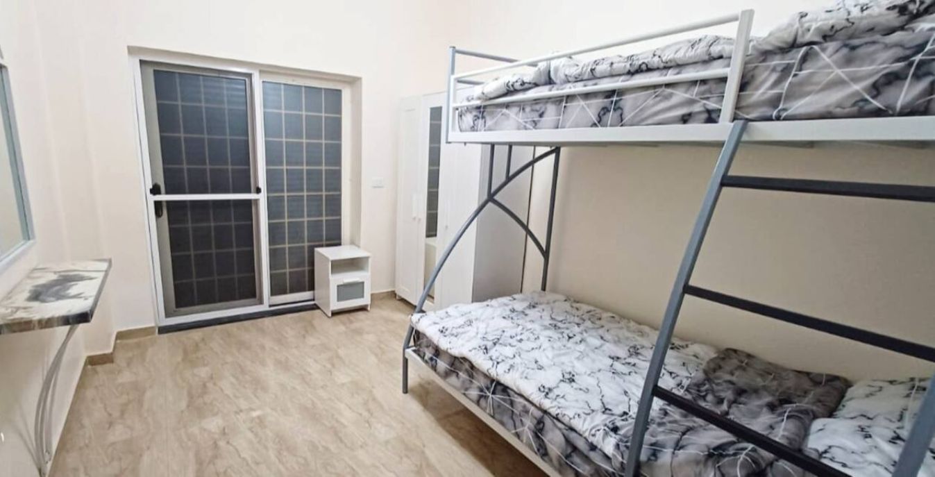 A dormitory room with bunk beds, a sliding glass door, a small table, and a window.
