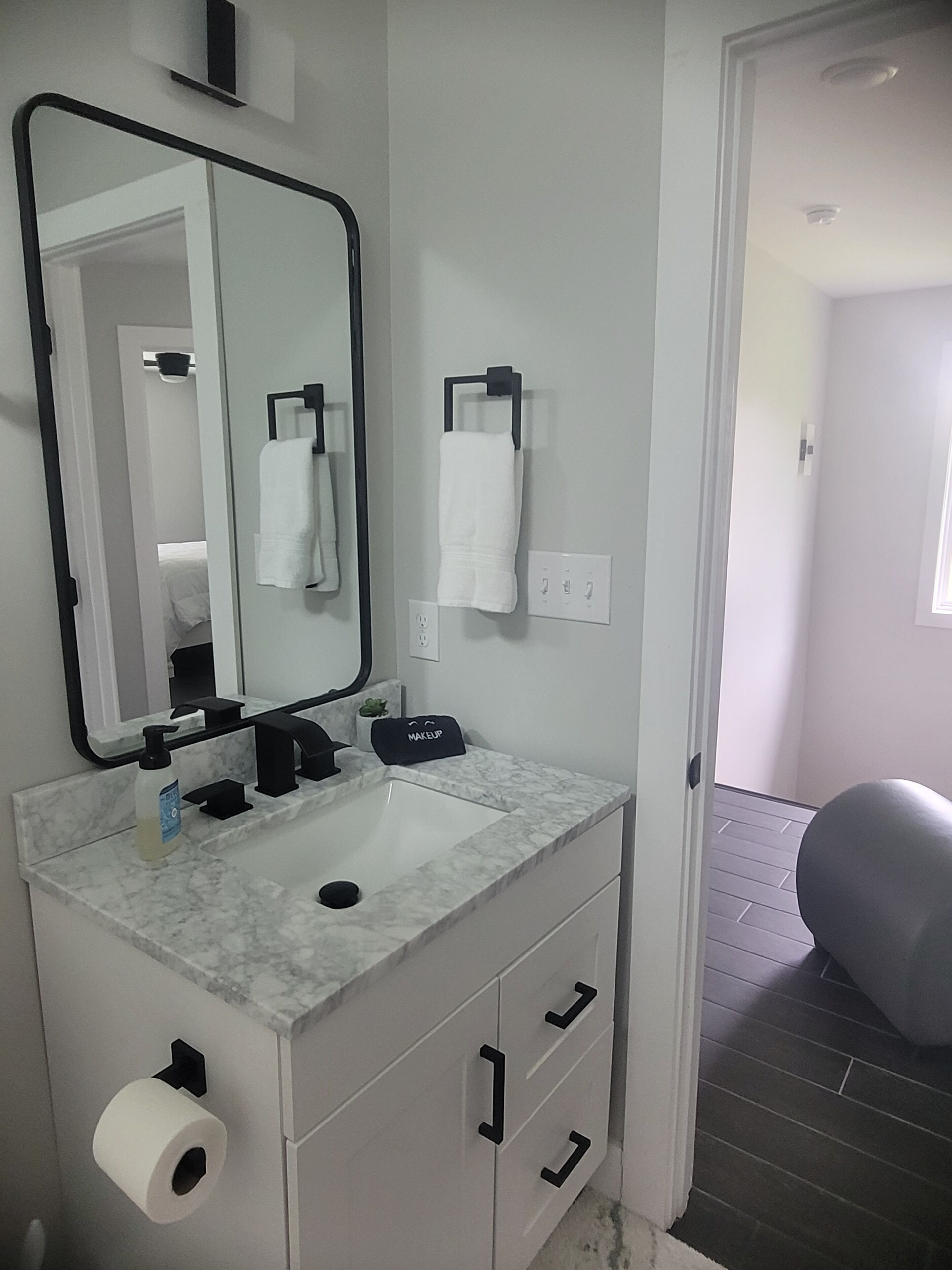 A bathroom with a marble sink, black faucet, a mirror, a towel rack, a makeup bag, and a toilet paper holder.
