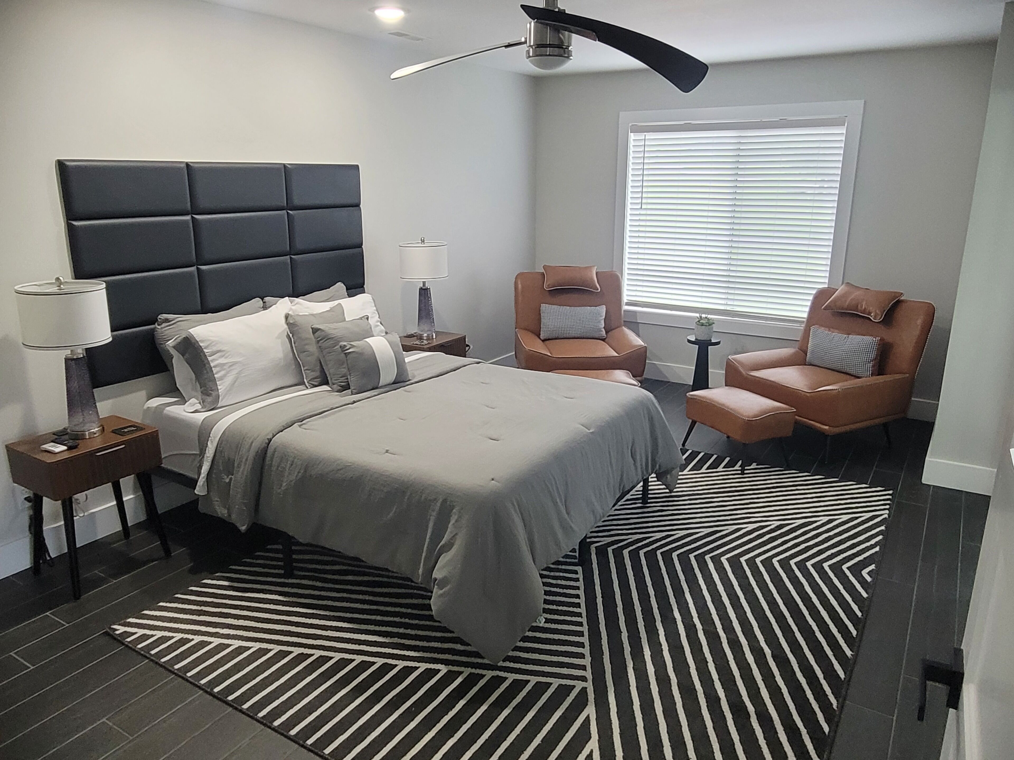 A modern bedroom with a large bed, two armchairs, a side table, a floor lamp, a ceiling fan, and a window with blinds.
