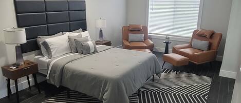 A modern bedroom with a large bed, two armchairs, a side table, a floor lamp, a ceiling fan, and a window with blinds.