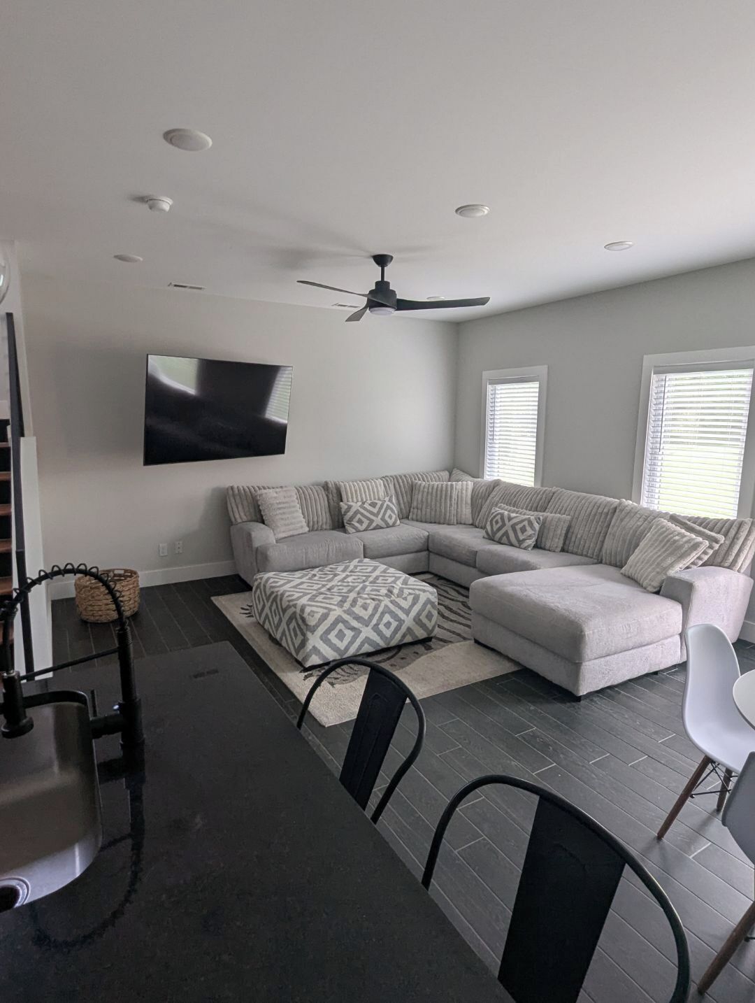 A modern living room with a sectional sofa, a patterned ottoman, a ceiling fan, and a flat-screen TV mounted on the wall.