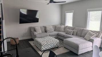 A modern living room with a sectional sofa, a patterned ottoman, a ceiling fan, and a flat-screen TV mounted on the wall.
