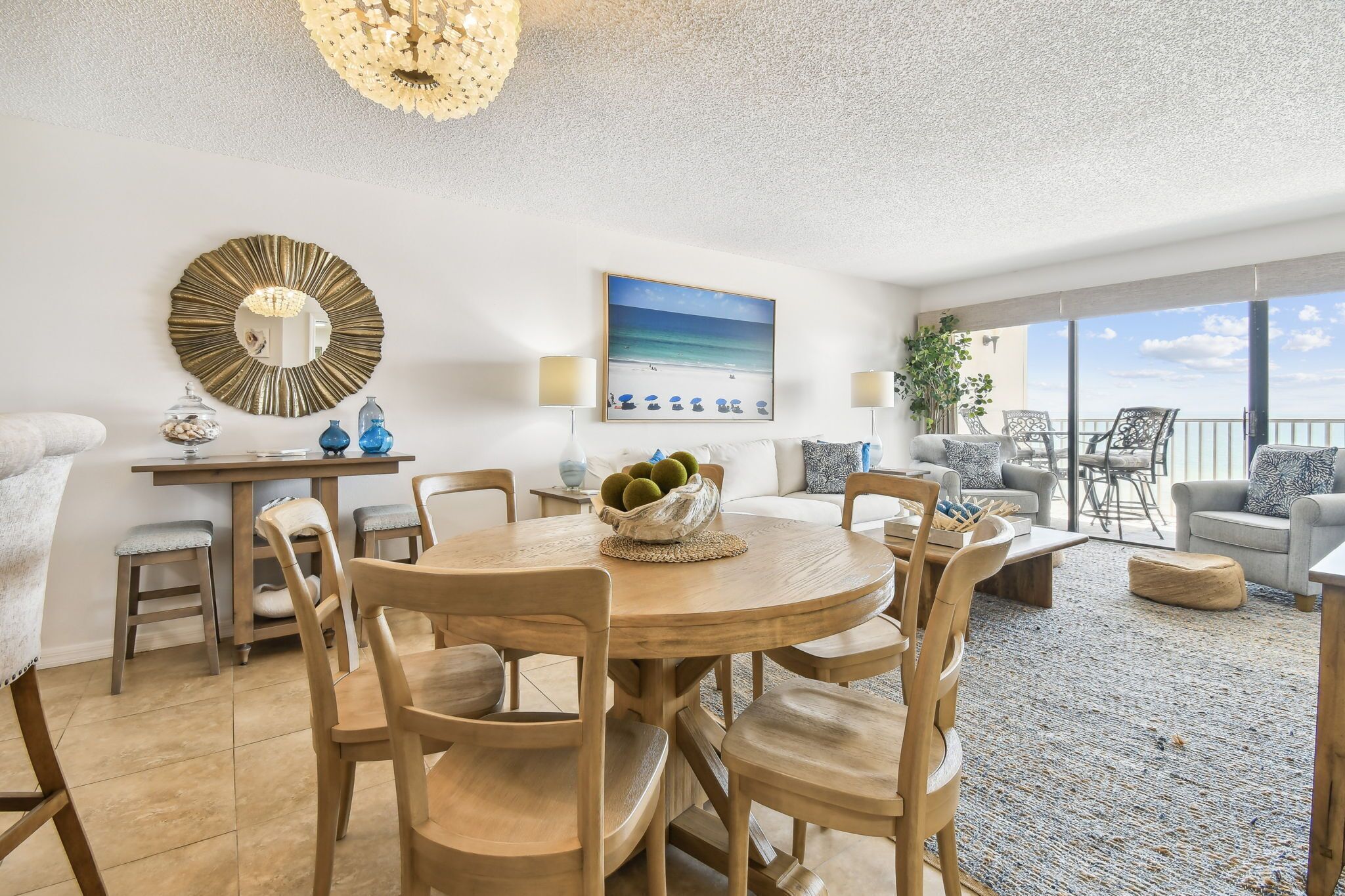 A spacious living area with a round wooden table, comfortable seating, a large mirror, and a view of the beach through large windows.