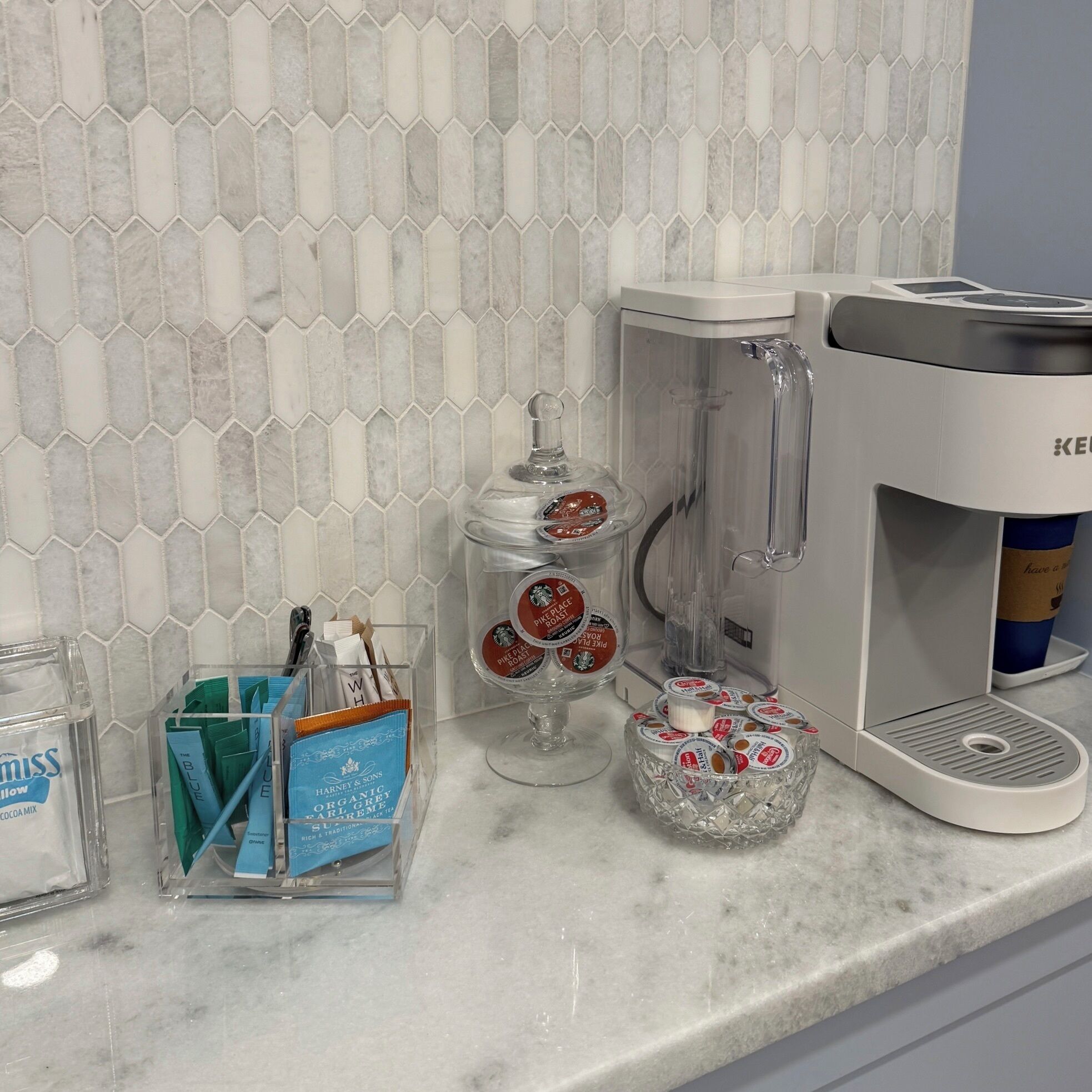 A kitchen counter with a coffee maker, a blender, a jar of coffee mix, and a container with tea bags.