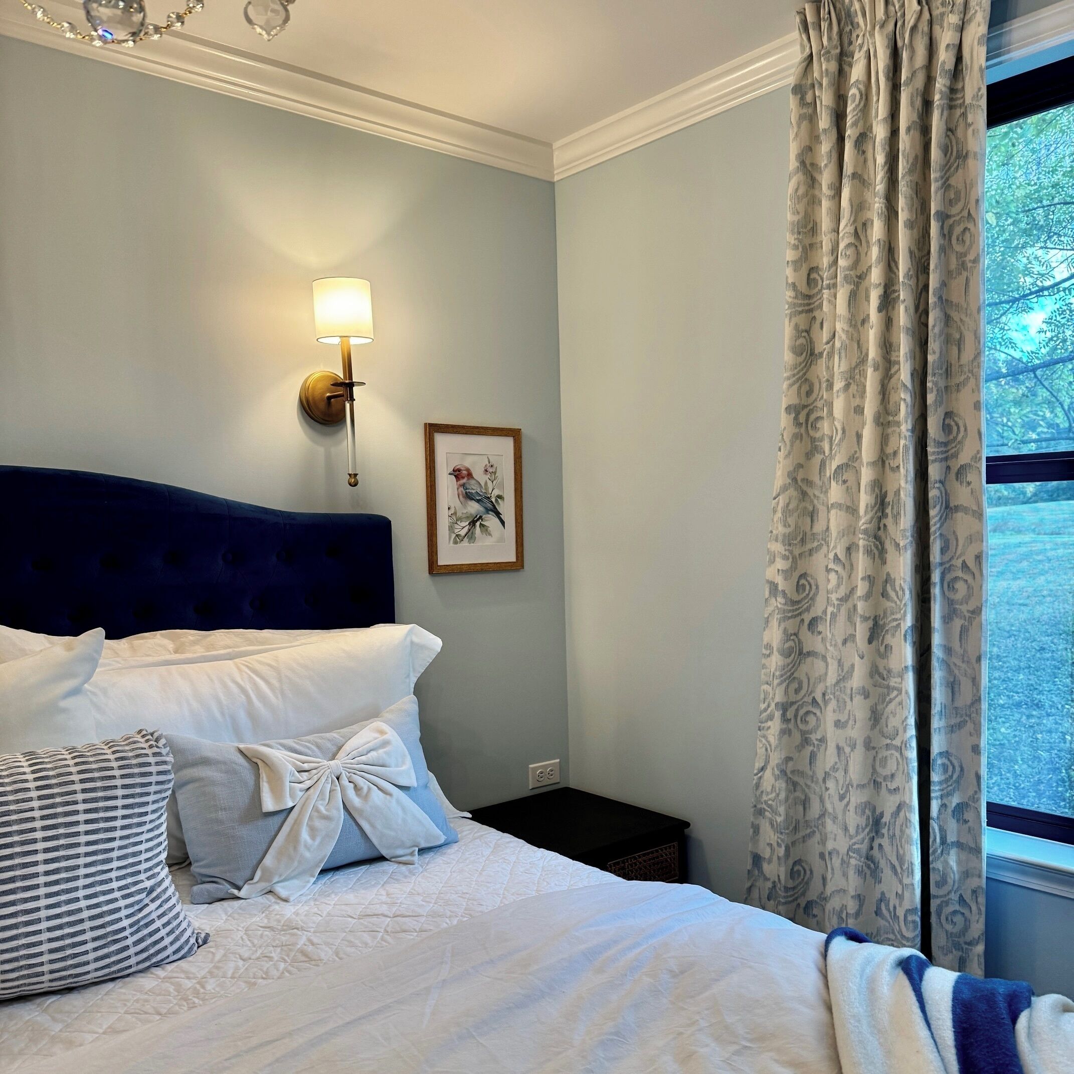 A bedroom with a bed, pillows, a wall-mounted lamp, a framed picture, and a window with curtains.