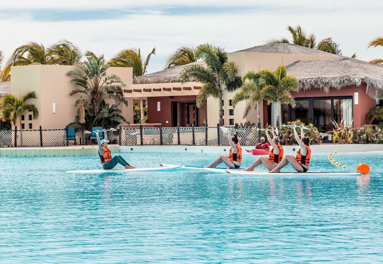 A swimming pool with people on surfboards, palm trees, and a building with a thatched roof in the background.