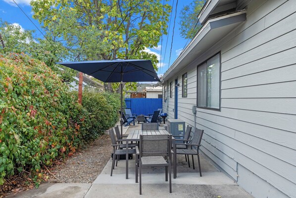 Une terrasse extérieure avec une table et des chaises, un parasol bleu et une haie.