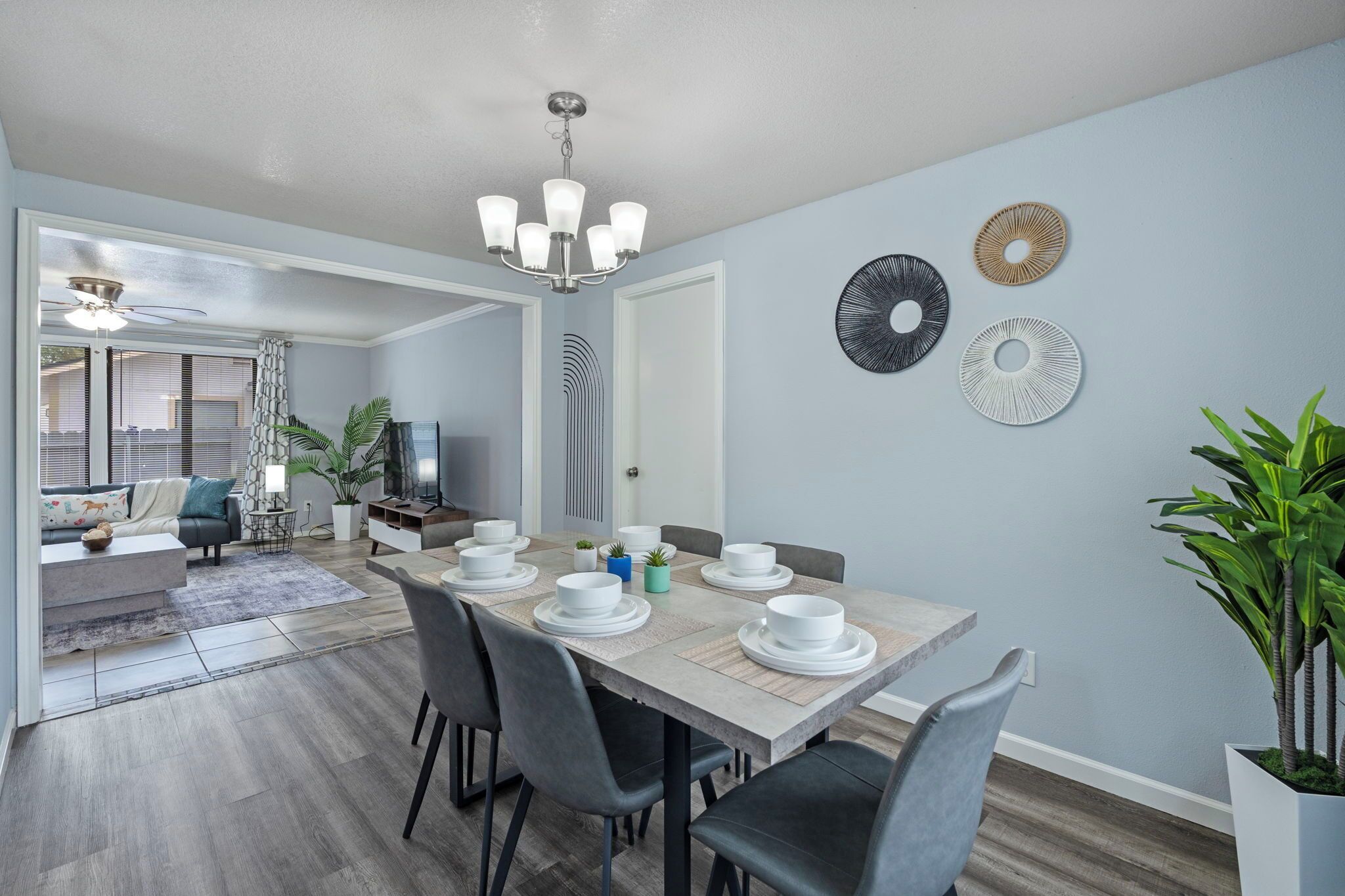 A dining area with a wooden table set for six, surrounded by grey chairs. There are decorative wall art pieces and a ceiling fan above the table.