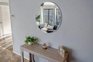 Un couloir avec un grand miroir rond qui reflète un salon, une console en bois sur laquelle sont posés une plante, un globe décoratif et un panneau.