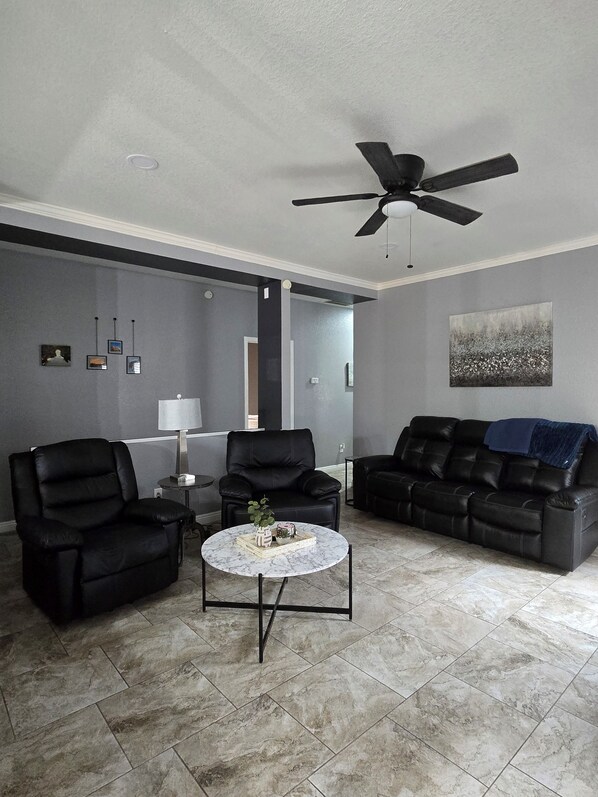 A modern living room with a ceiling fan, two black leather sofas, a marble coffee table, and a grey tiled floor.