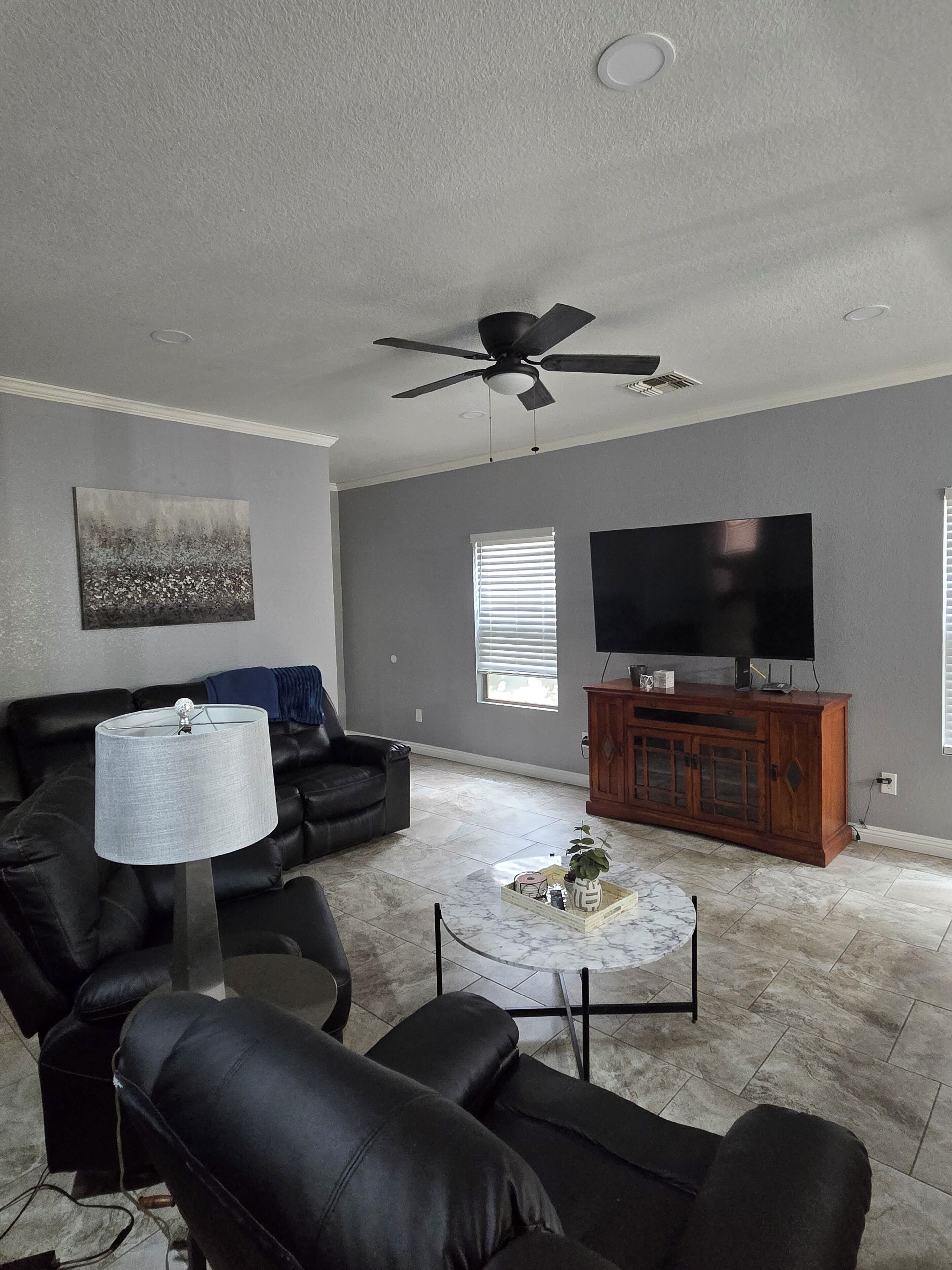 A living room with a black leather sofa, a coffee table, a TV on a wooden cabinet, and a ceiling fan.