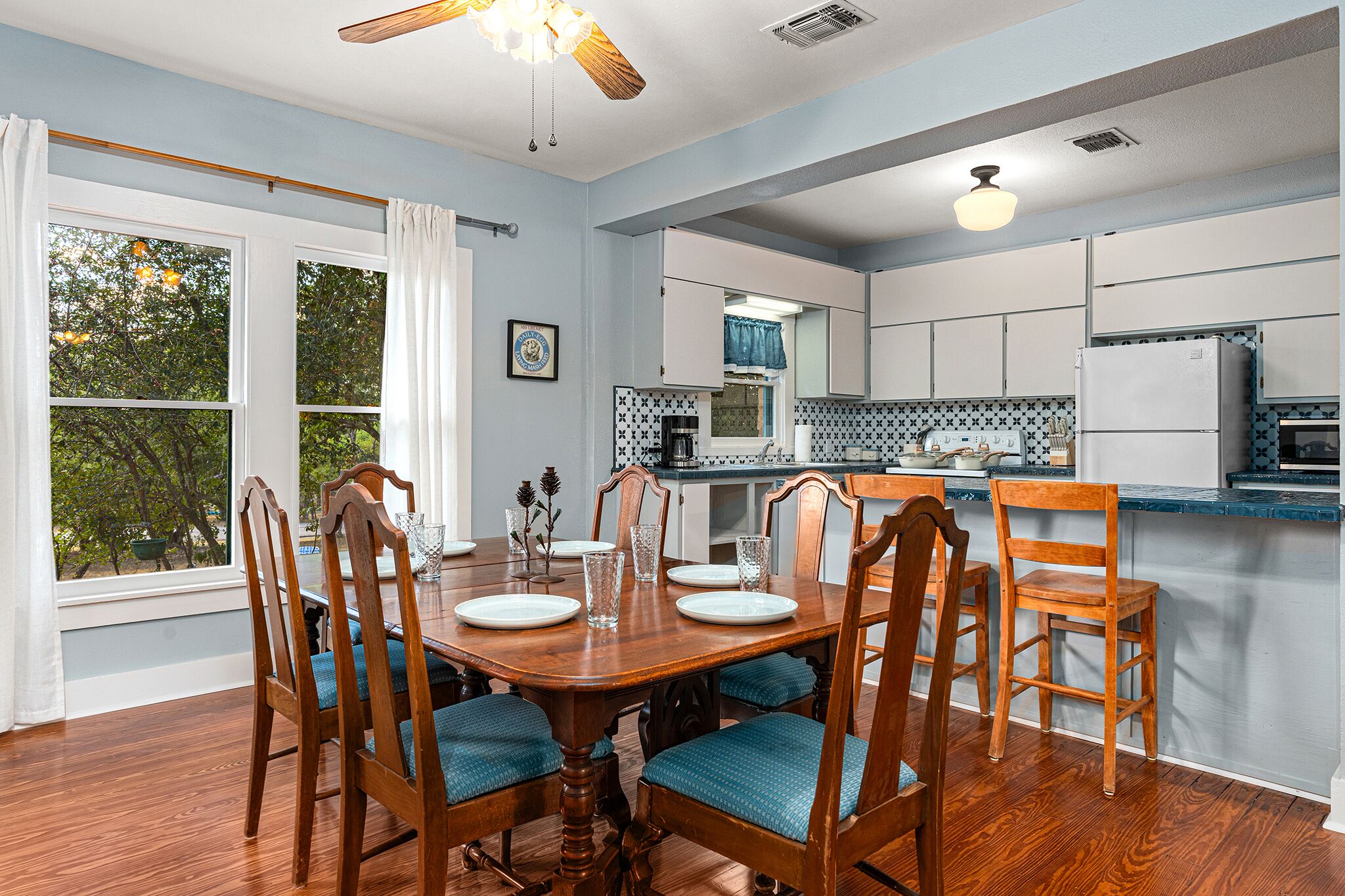 A dining area with a wooden table and chairs, a kitchen with white cabinets, and a view of trees through large windows.