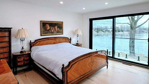 Een slaapkamer met een houten bed, twee nachtkastjes met lampen, een schilderij aan de muur en een groot raam met uitzicht op een wateroppervlak.