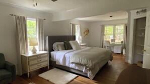 Une chambre avec un grand lit, des tables de chevet, un bureau et une chaise. Il y a des fenêtres avec des rideaux, un ventilateur de plafond et une porte qui donne sur une autre pièce.