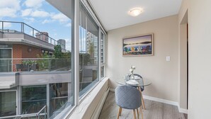 A balcony with a glass railing, a dining table with chairs, a painting on the wall, and a view of buildings and trees. - Downtown Vancouver Condo, Pool, Gym, Skytrain (Vancouver)