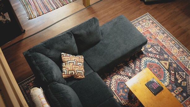 A modern living room with a dark grey sofa, a patterned throw pillow, and a wooden coffee table with a book. A colorful rug covers the wooden floor, and a patterned rug is visible in the background.