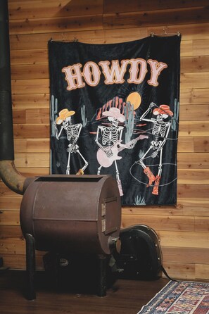 A rustic room with a wooden wall, a black tapestry with skeletons and the word "HOWDY," a large brown barrel, and a black vacuum cleaner.