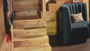 Una escalera de madera que baja a un nivel inferior, un sillón oscuro con cojín, y un tapiz de colores.