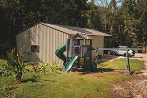 A playground with a slide and a small building, surrounded by trees and a parked car.