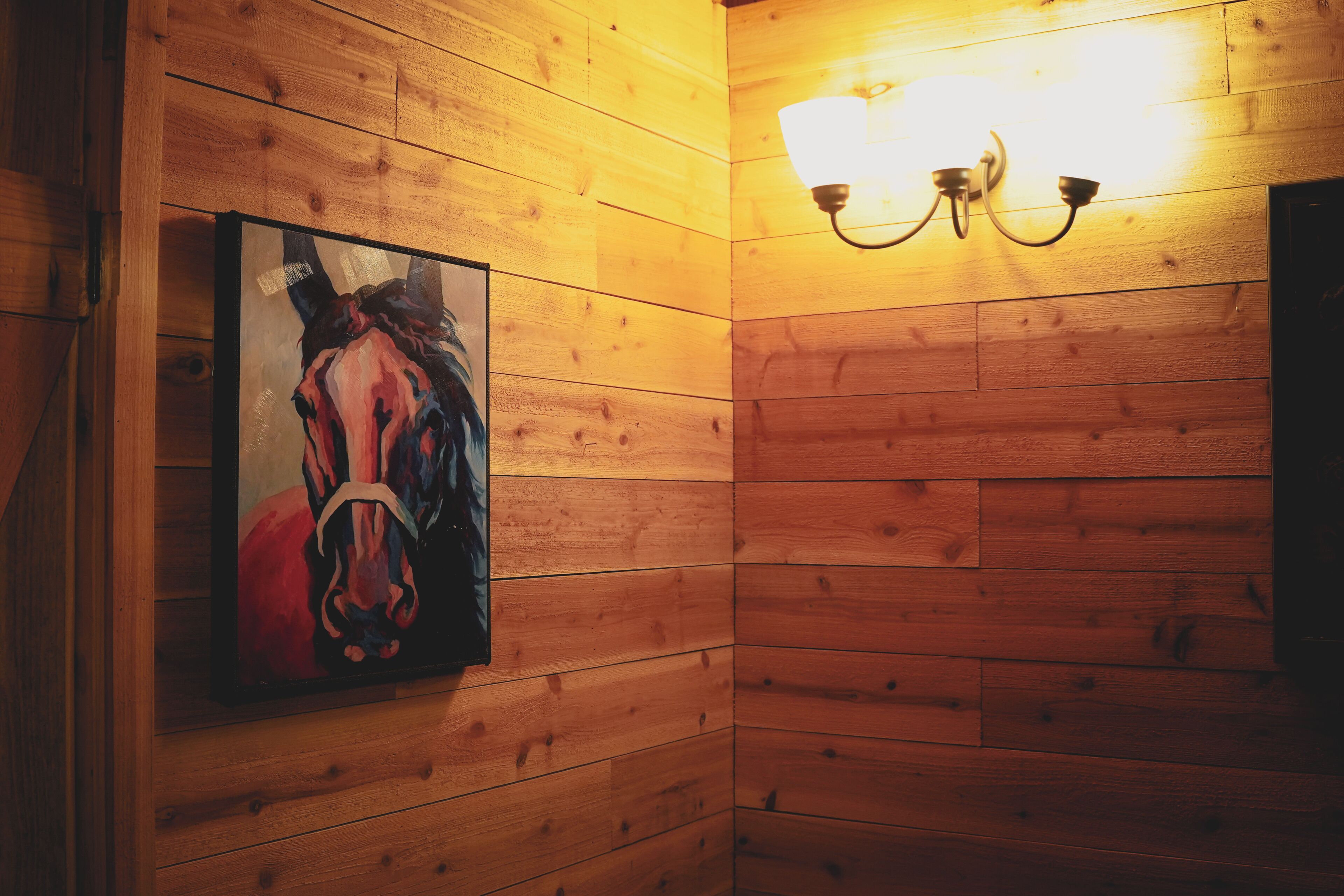 A wooden wall with a framed painting of a horse and a wall-mounted light fixture.
