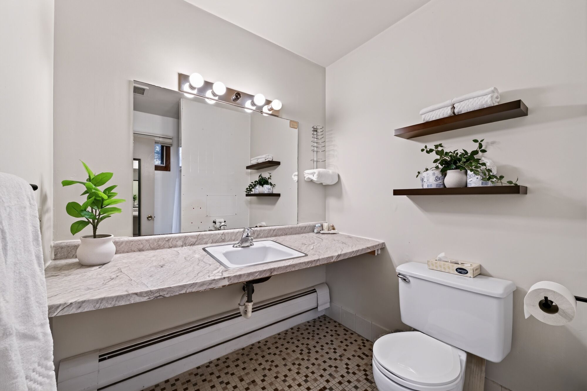 A bathroom with a marble sink, a toilet, a mirror with lights, and shelves with towels and plants.