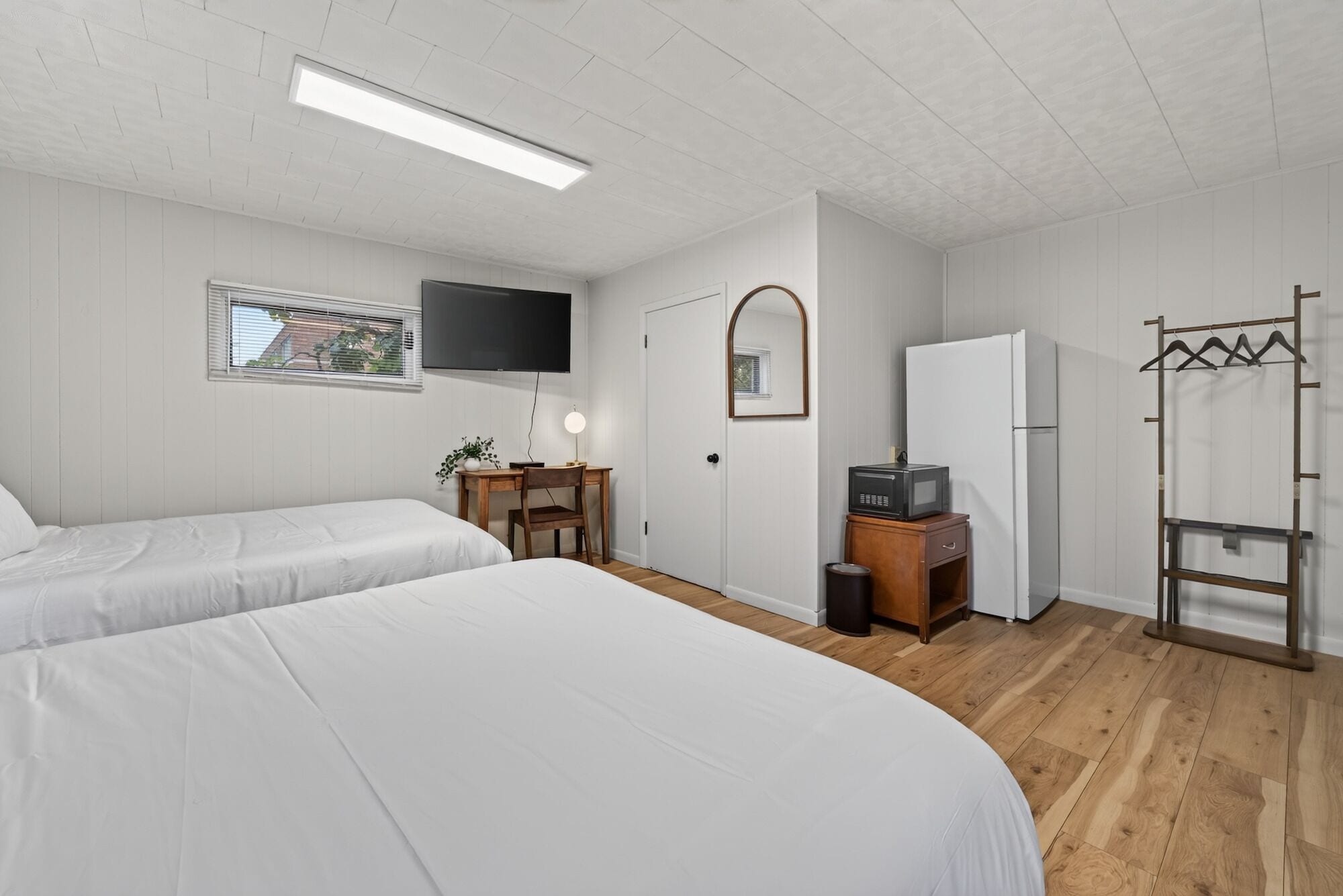 Une chambre avec deux lits, un bureau, une télévision, un miroir, un réfrigérateur et un porte-manteau.