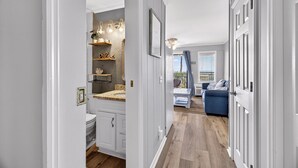 Un couloir mène à une salle de bain équipée de toilettes, d’un lavabo et d’étagères où sont disposés des objets décoratifs. On aperçoit un canapé bleu et une fenêtre avec des rideaux au fond.