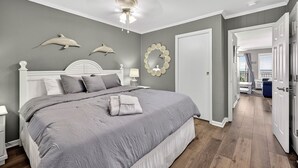 Une chambre avec un grand lit, un ventilateur de plafond et une porte donnant sur une autre pièce.