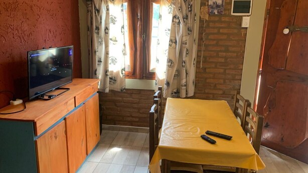 Quarto com televisão sobre um armário de madeira, uma mesa de jantar com cadeiras e uma janela com cortinas.