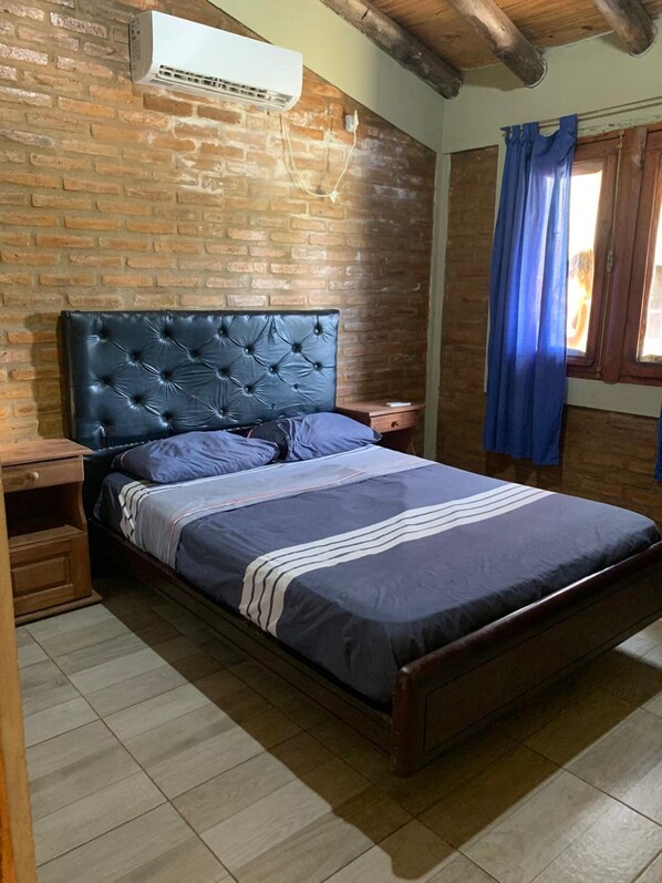 A bedroom with a brick wall, a wooden ceiling, a bed with a tufted headboard, a nightstand, and a window with blue curtains.