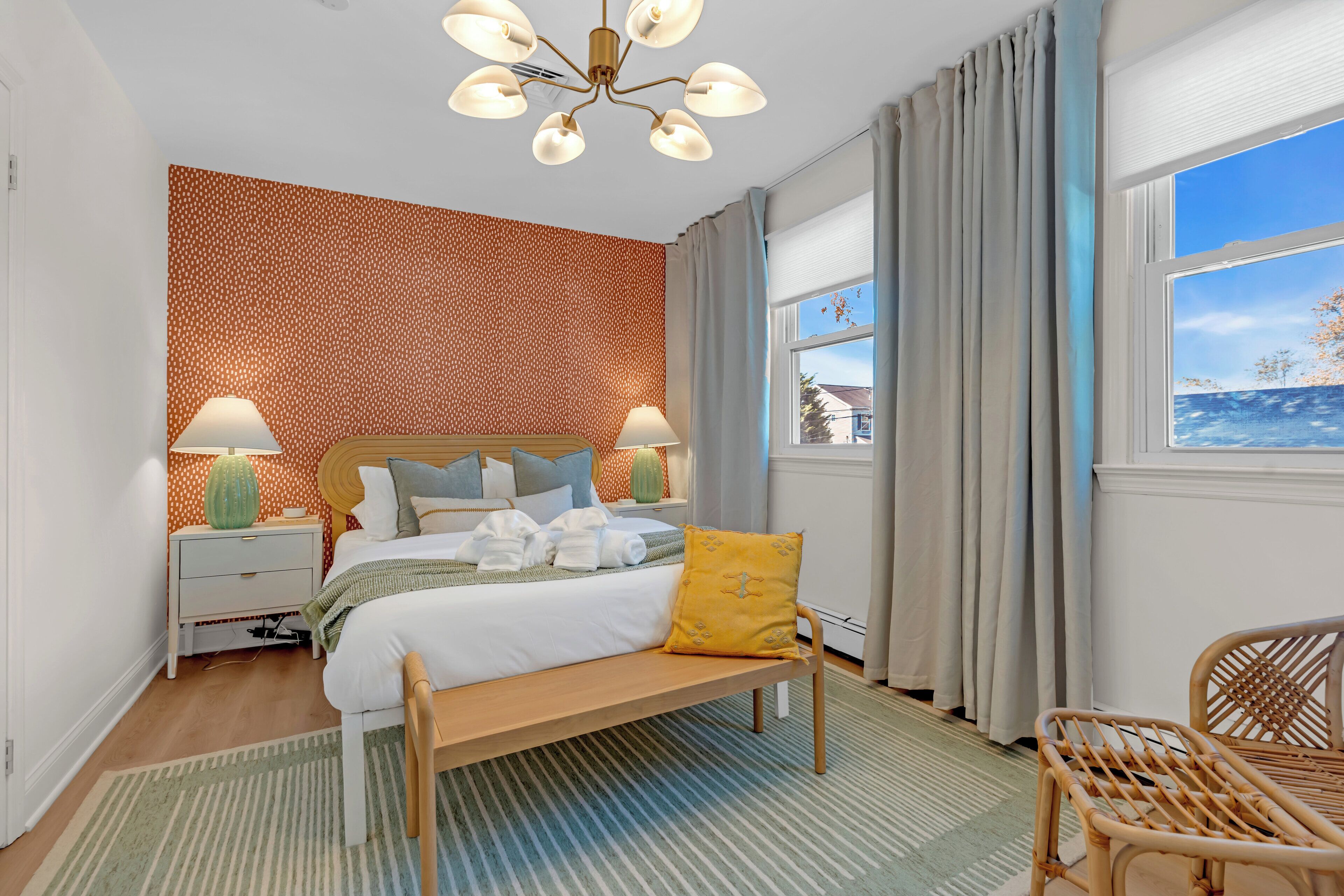 Een slaapkamer met een bed, bedlampen, een bankje, een stoel, een raam met gordijnen en een behang met patroon.