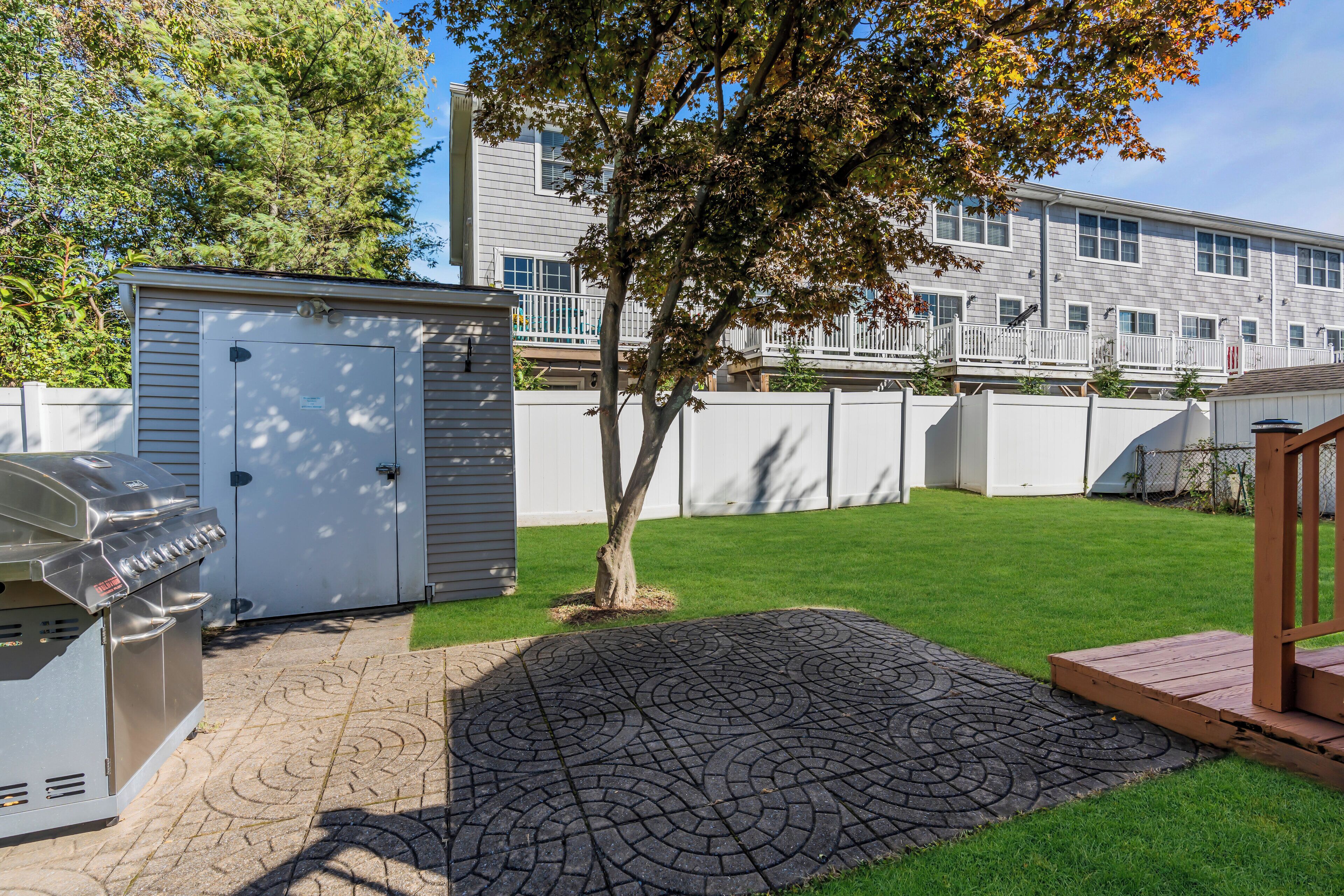 Ein Garten mit Grill, Gerätehaus, Baum und gepflasterter Terrasse.