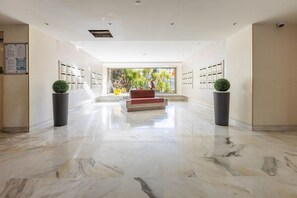 A spacious lobby with a marble floor, a central red bench, and potted plants.