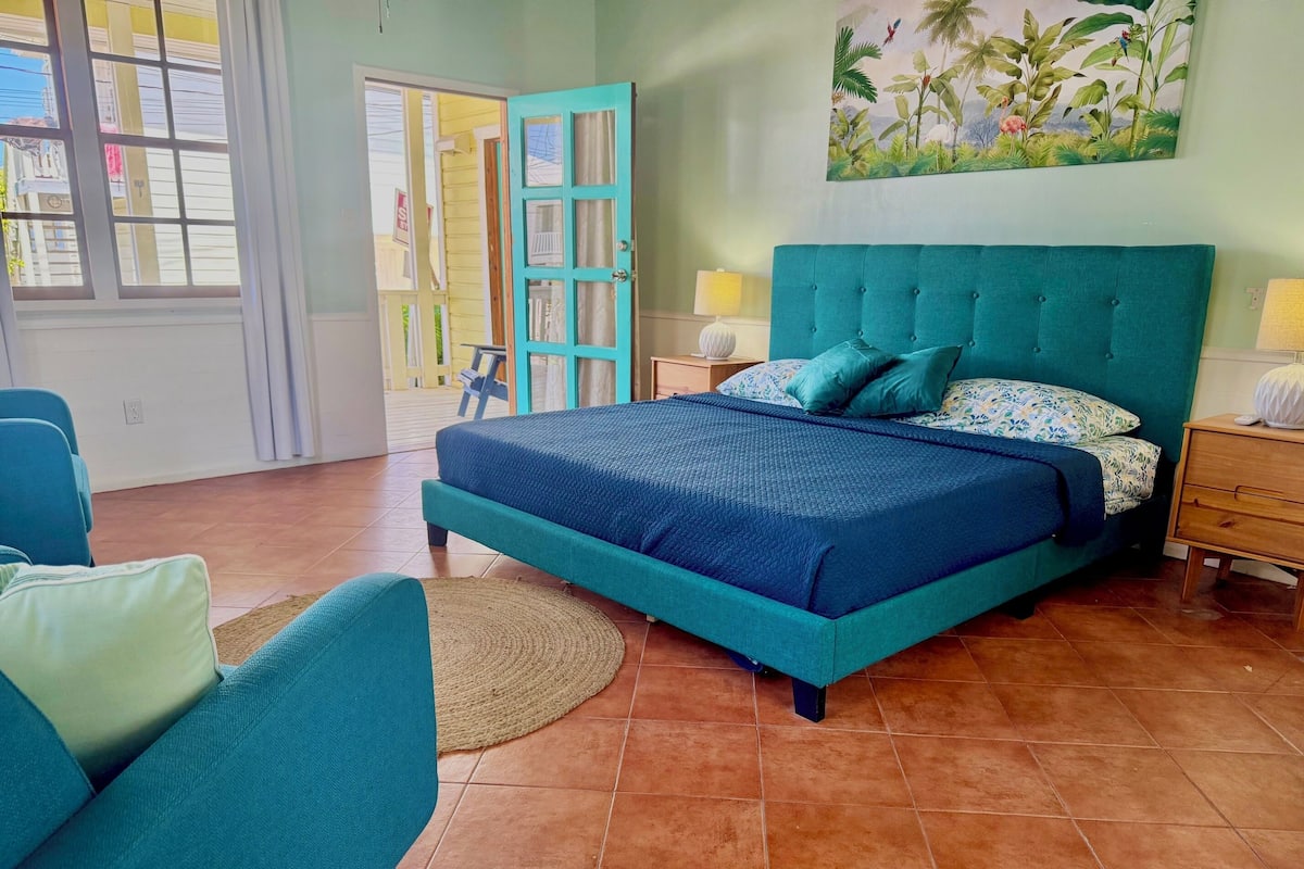 Ein Schlafzimmer mit einem türkisfarbenen Bett, einem Sessel, einem Nachttisch und einem Bild von tropischen Pflanzen.