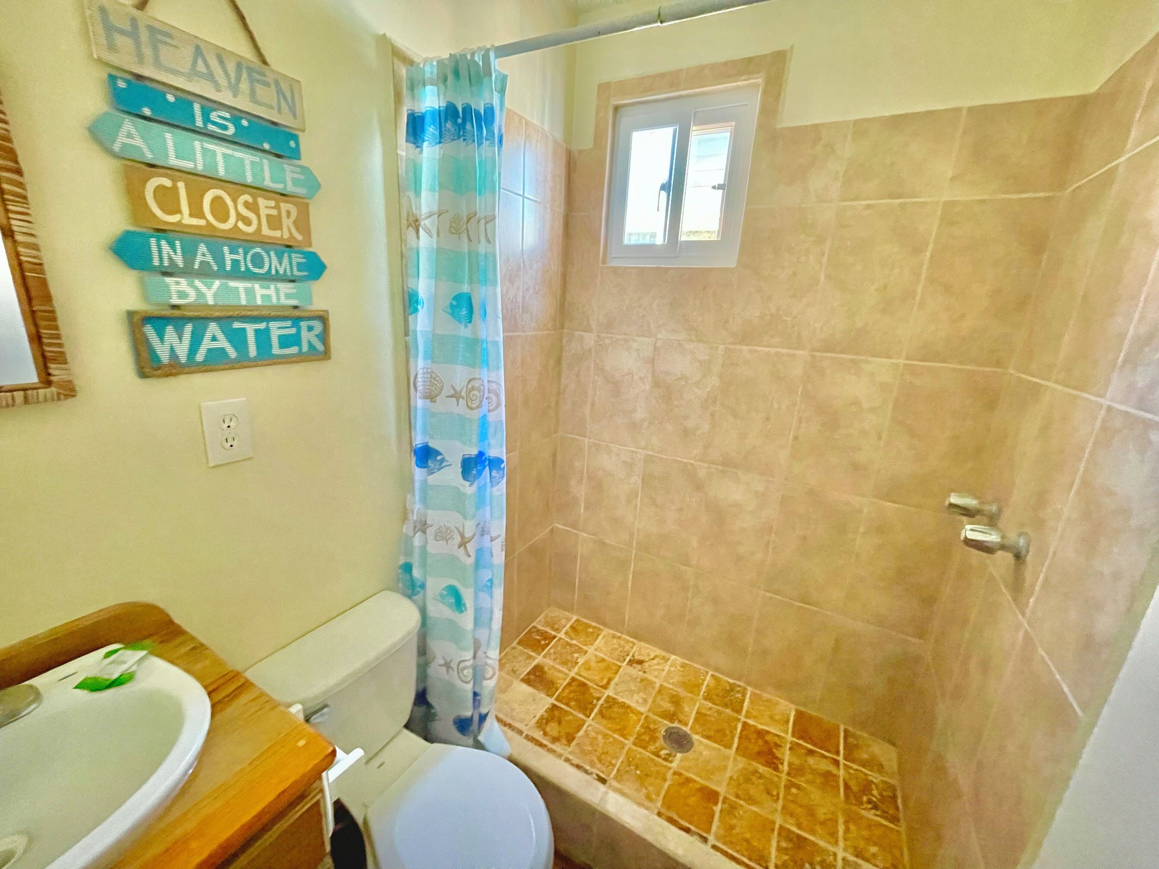 A bathroom with a shower, toilet, and sink. There are decorative signs on the wall and a window above the shower.