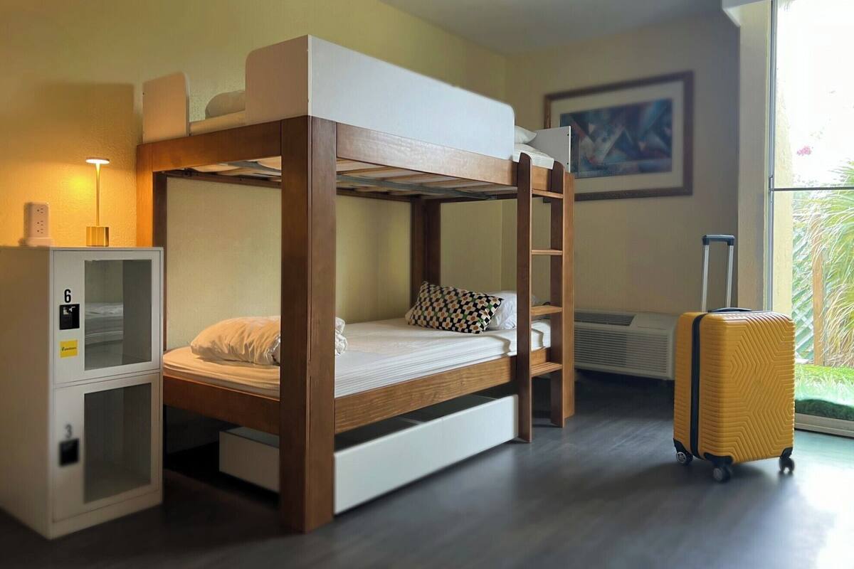 A bunk bed room with a desk, a suitcase, and a window with a view of greenery.
