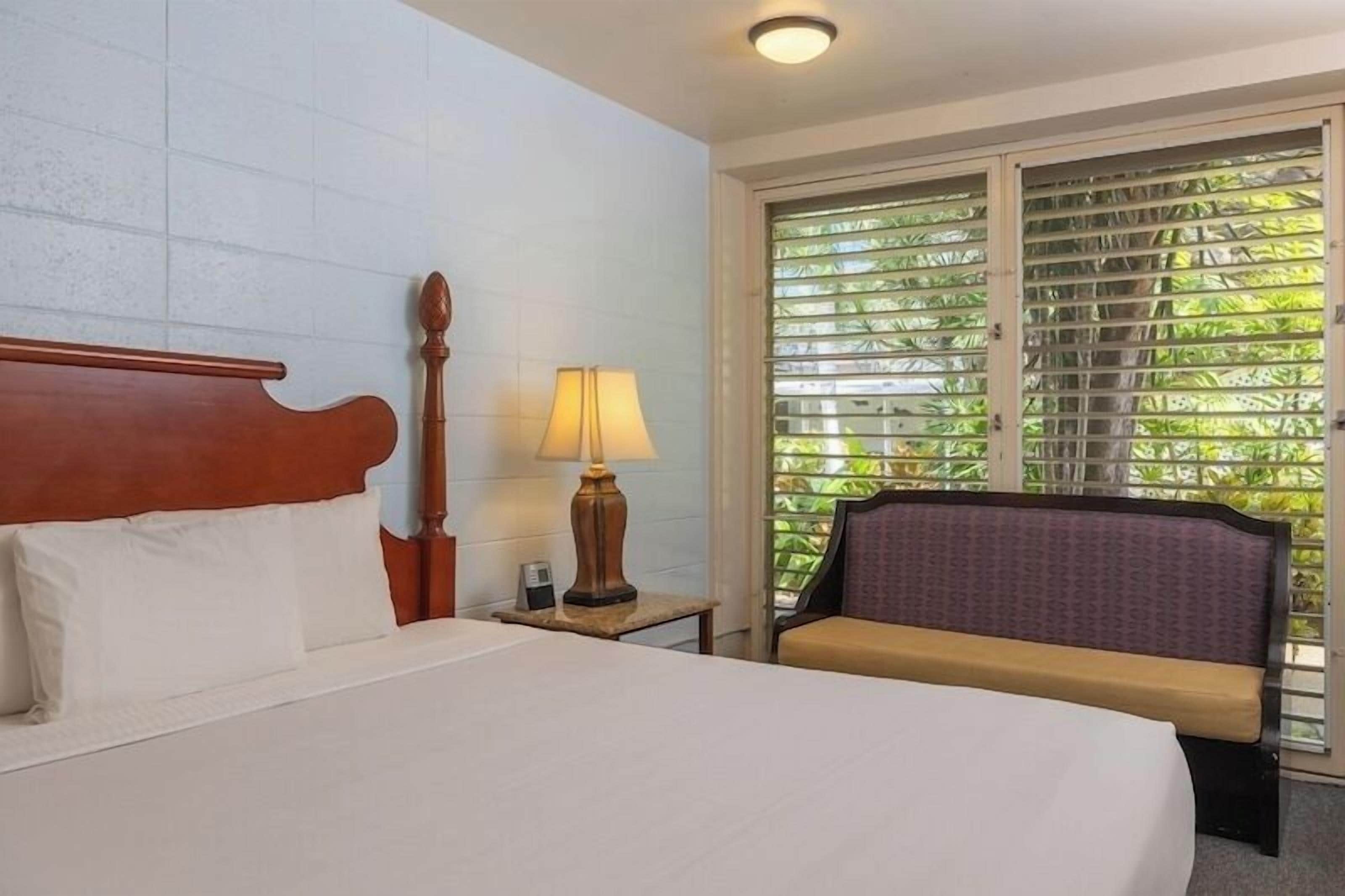 A hotel room with a bed, a nightstand, a lamp, a chair, and a window with blinds.