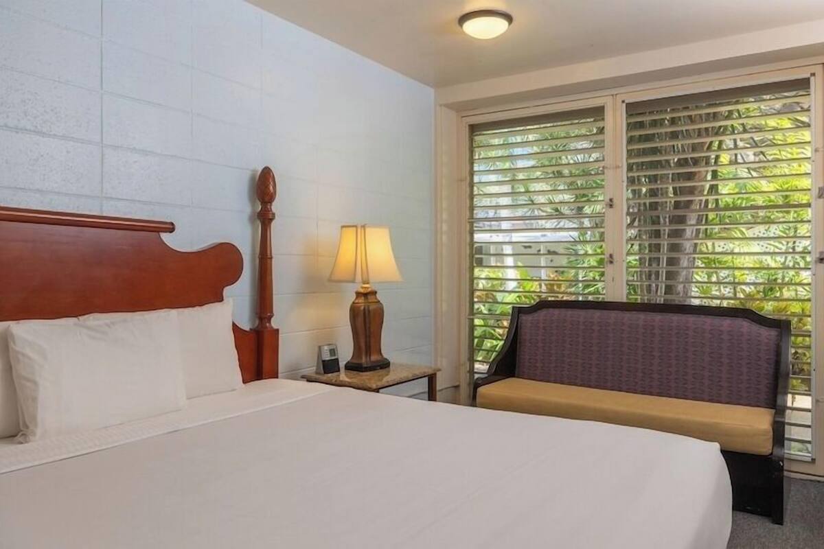A hotel room with a bed, a nightstand, a lamp, a chair, and a window with blinds.
