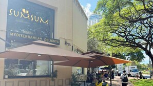Un espacio al aire libre con sillas y mesas de mimbre, un cartel que dice 'Suxisux Mediterranean Delights' y un edificio al fondo.