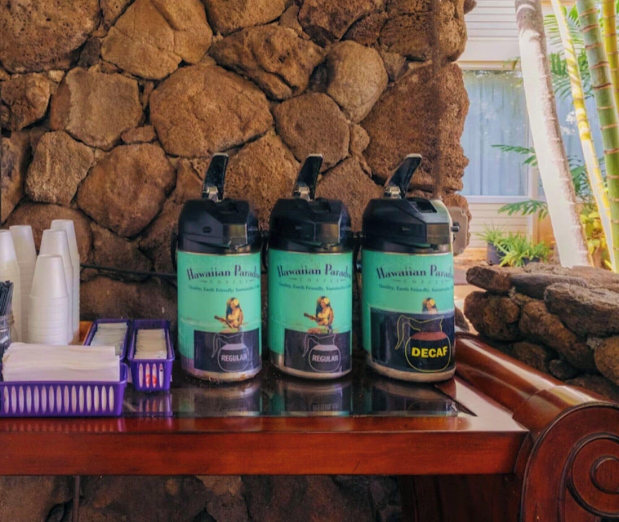 A table with three Hawaiian Paradise coffee containers, a stack of white cups, and a purple basket with napkins.