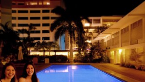 Una piscina con dos personas sentadas al borde, rodeada de palmeras y edificios al fondo.