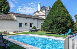 Une piscine avec un espace patio, un grand arbre à feuilles persistantes et un bâtiment doté d’un toit à pignon.
