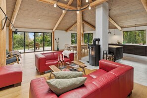 Un vaste espace de vie dotĂ© dâun plafond haut, de poutres en bois, dâune cheminĂ©e et de grandes fenĂȘtres offrant une vue sur lâextĂ©rieur.