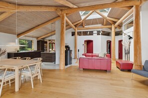 Ein geräumiger Innenraum mit hoher Holzbalkendecke, einer Essecke mit Holztisch und Stühlen, einem roten Sofa und einem grauen Sessel.
