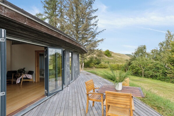 Eine Holzterrasse mit Tisch und Stühlen, eine Glastür zu einem Innenraum mit Sessel und Tisch sowie Blick auf eine grüne Landschaft mit Bäumen.