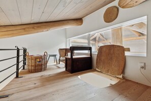 Une chambre situĂ©e dans un espace de type « loft », avec un plafond en pente, des poutres en bois, une grande fenĂȘtre, une chaise en bois, un panier en osier et une petite table.