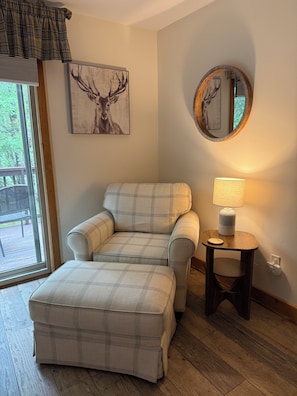 A cozy room with a plaid armchair and ottoman, a round mirror, and a painting of deer antlers.