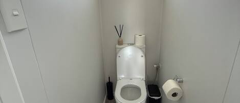 Een smalle badkamer met een toilet, een klein raam en een prullenbak.