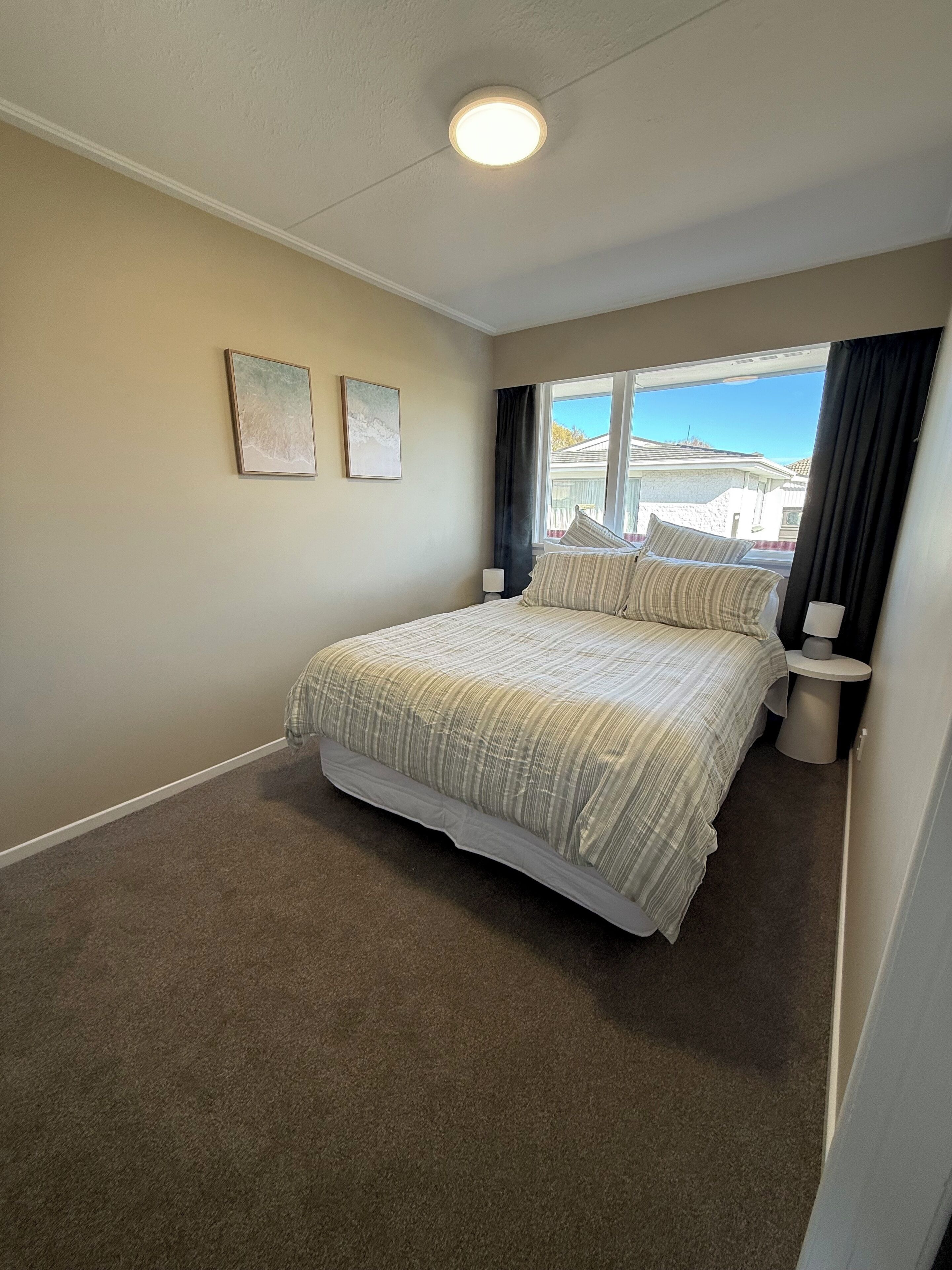 Un dormitorio con una cama grande, dos cuadros en la pared, un ventanal con cortinas y vistas a casas.